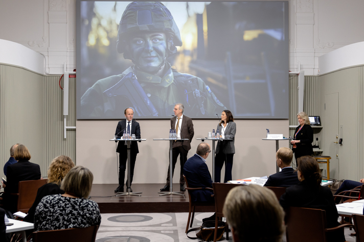 Tre personer står på en scen och pratar. I bakgrunden syns en bild av en värnpliktig.