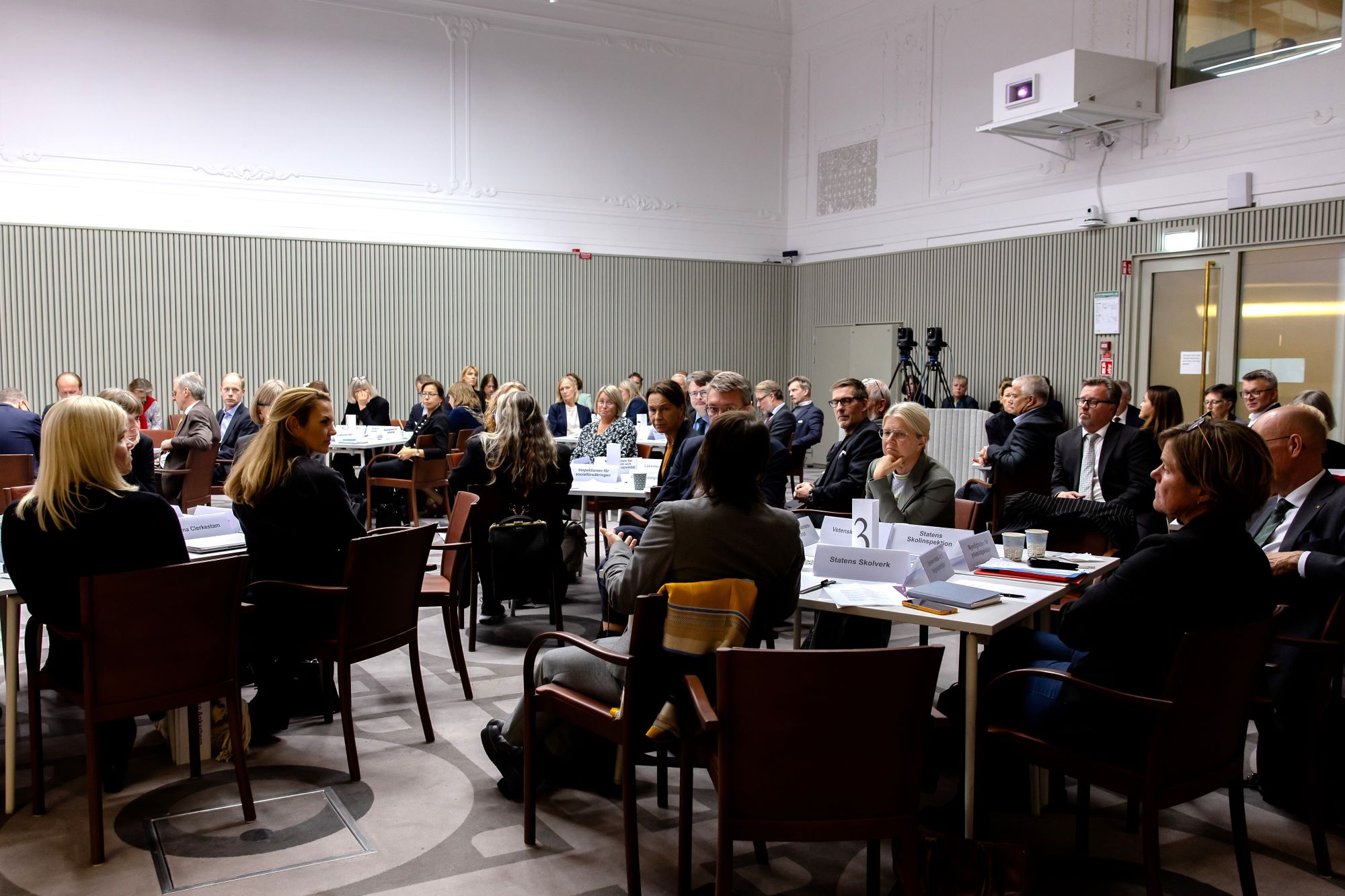 Ett stort rum med flera bord och personer som sitter runt borden och samtalar.