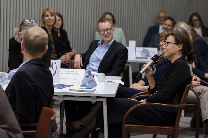 Flera personer sitter runt ett bord. En håller i en mikrofon och pratar.