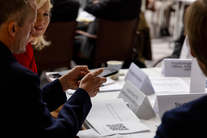 Några personer sitter runt ett bord och pratar. En håller i en mobil.