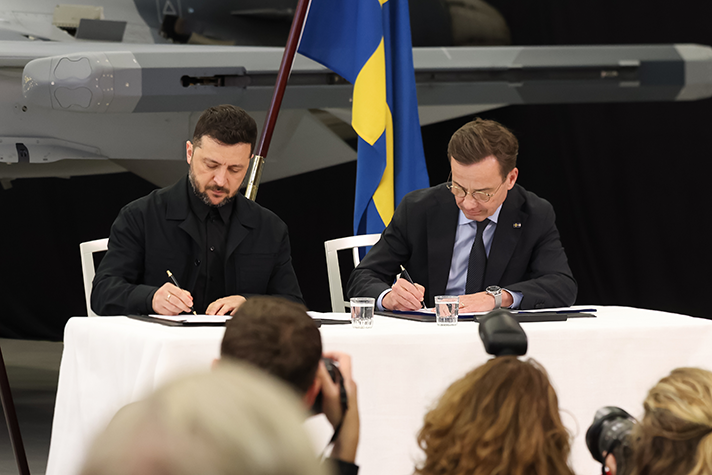 Volodymyr Zelensky och Ulf Kristersson sitter vid ett bord och signerar varsitt papper. I förgrunden syns journalister.