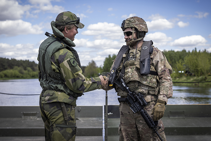 En svensk soldat och en amerikansk soldat i uniform står på en bro och skakar hand under en militär övning.