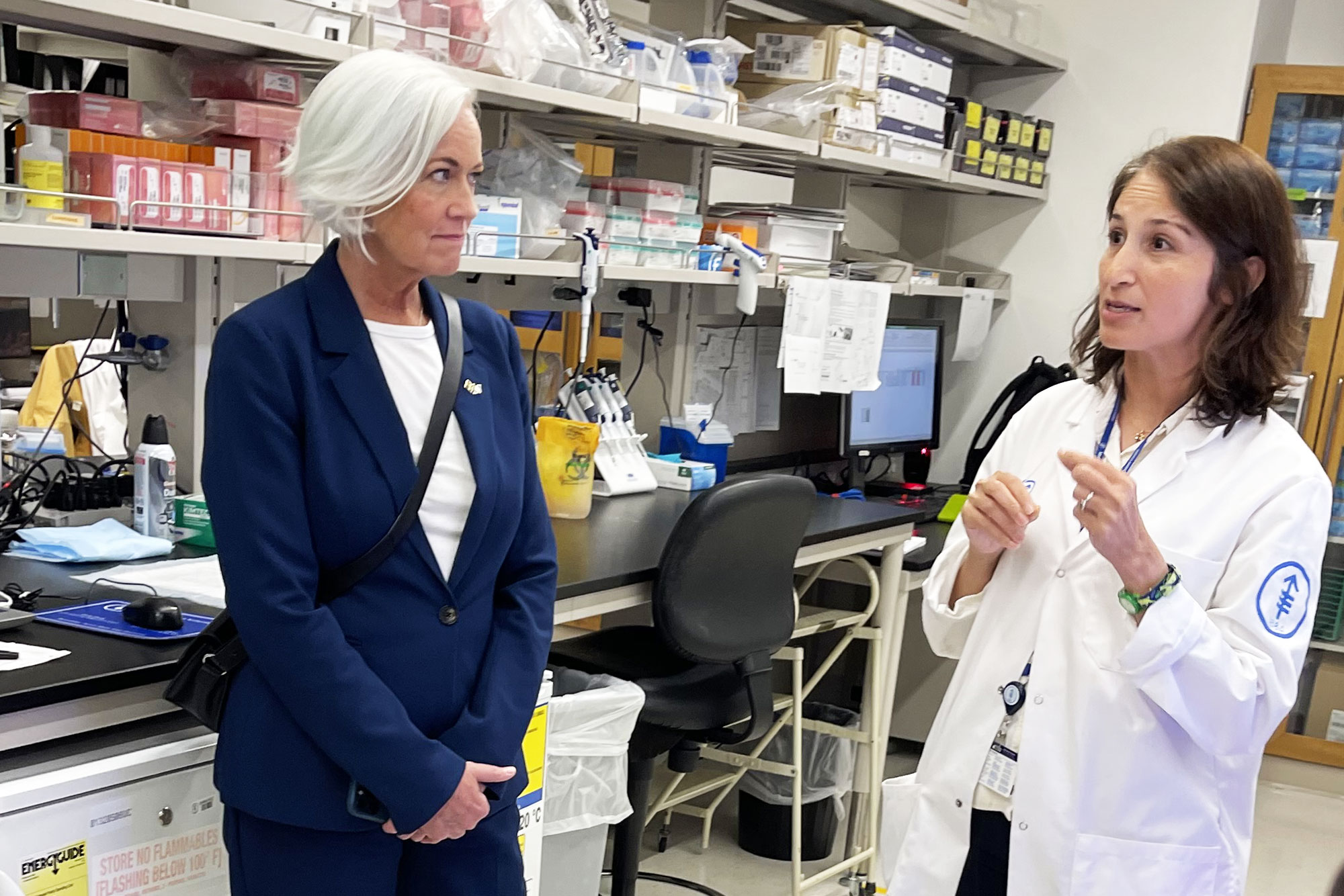 Acko Ankarberg Johansson lyssnar på en kvinnlig läkare i vit rock. De befinner sig en laboratoriemiljö.