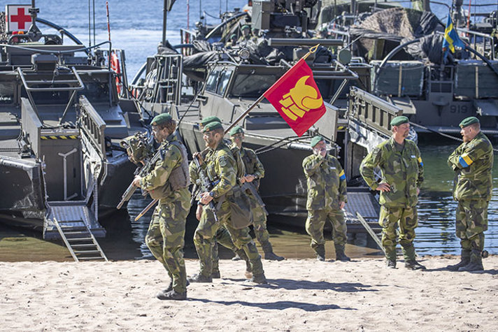 Amfibiesoldater marscherar med en röd flagga, prydd med ett huvud med vikingahjälm, på en strand längsmed uppställda stridsbåtar