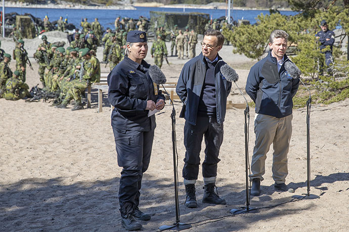 Pressträff på en sandstrand med amfibiesoldater i bakgrunden. Marinchefen Ewa Skoog Haslum står och pratar. Th om henne står statsminister Ulf Kristersson och försvarsminister Pål Jonson