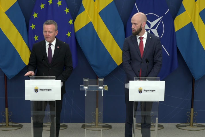 De två medverkande står vid var sin talarpulpet. Det är fanor i fonden.