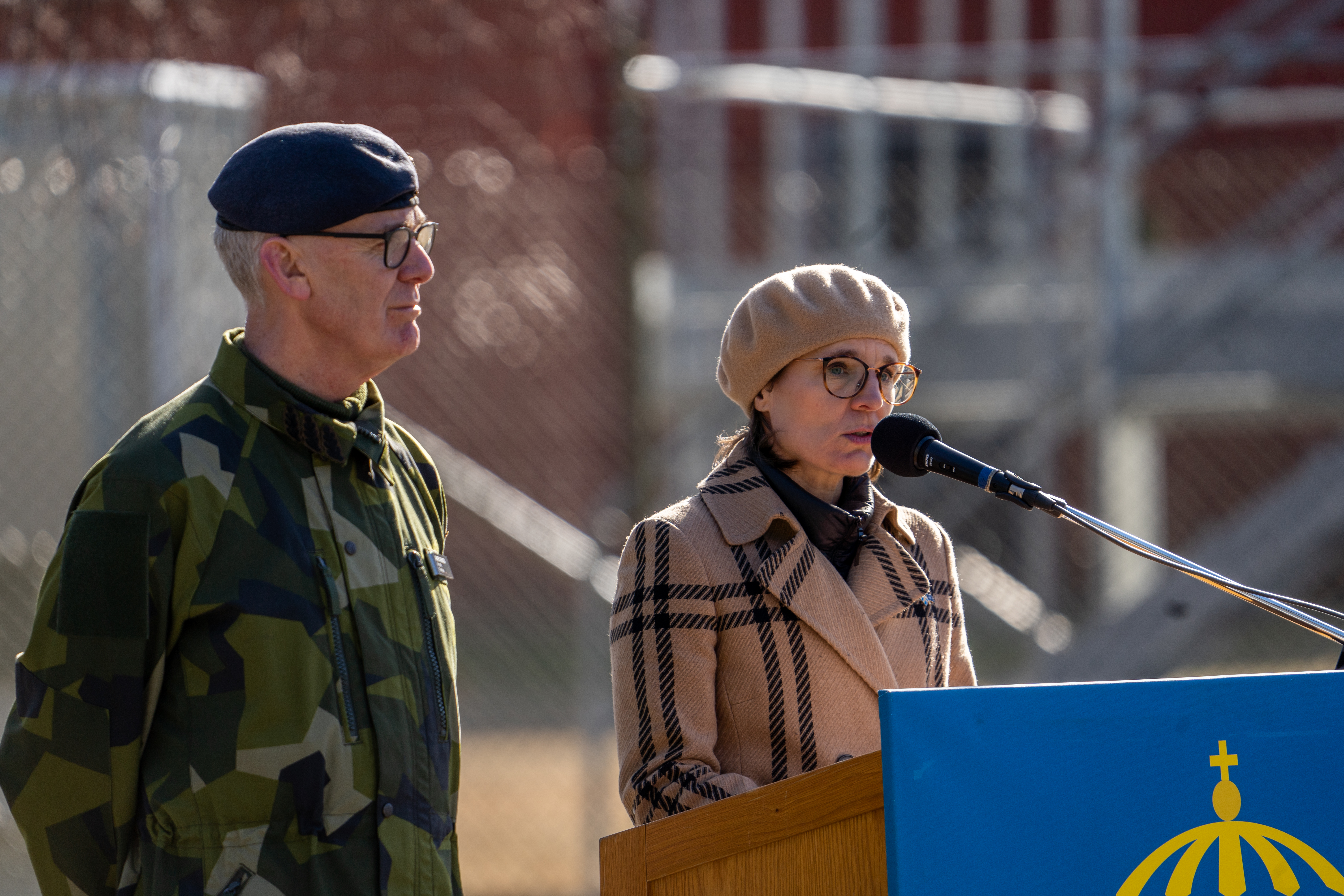 Jessika stående utomhus i kappa och basker vid en talarstol.