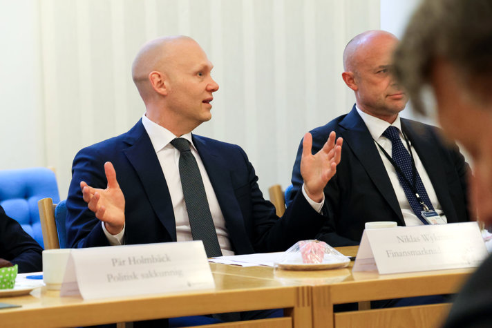 Man sitter vid ett bord och pratar och gestikulerar med händerna. Man anar en ytterligare person bredvid honom, och en annan i förgrunden.