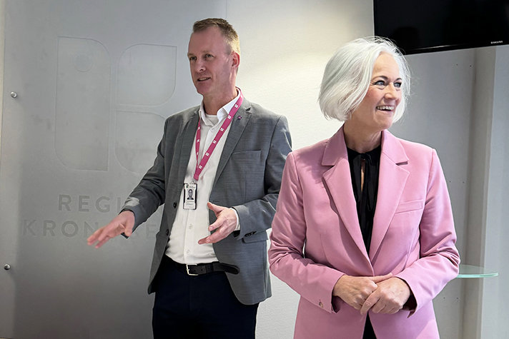 En man och en kvinna står framför en grå vägg där det står Region Kronoberg