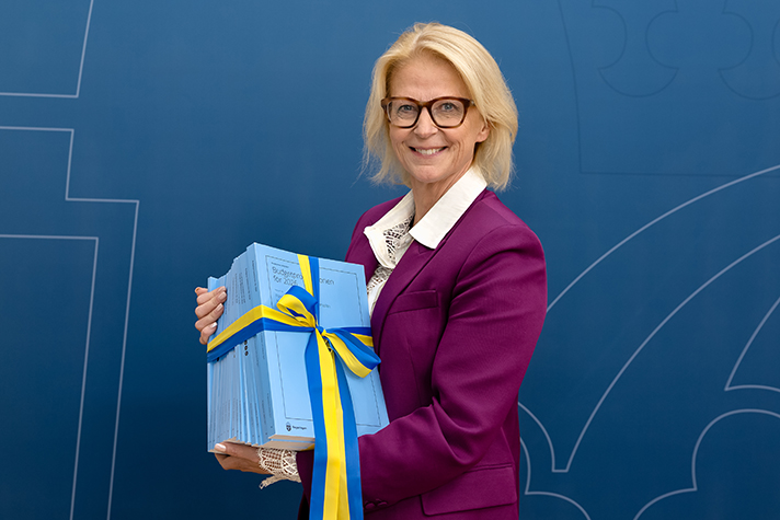 Finansminister i lila kostym med upptryckt budgetproposition i handen. Hon står framför Regeringskansliets backdrop.