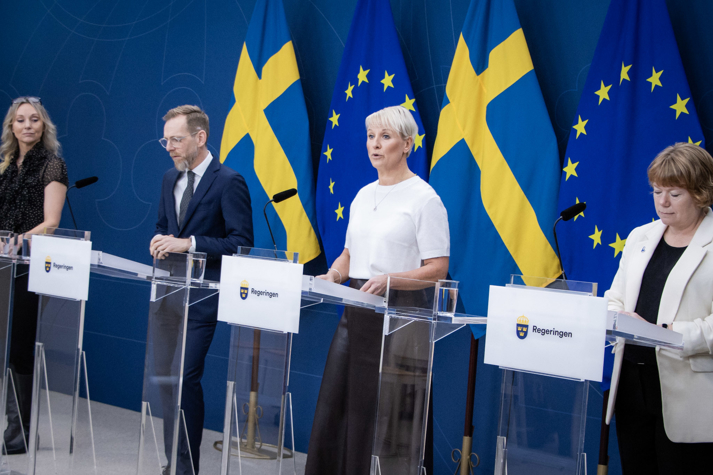 Anna Tenje och Jakob Forssmed står bakom var sin talarstol i reegeringens pressrum. I bakgrunden syns svenska flaggor och EU-flaggor.