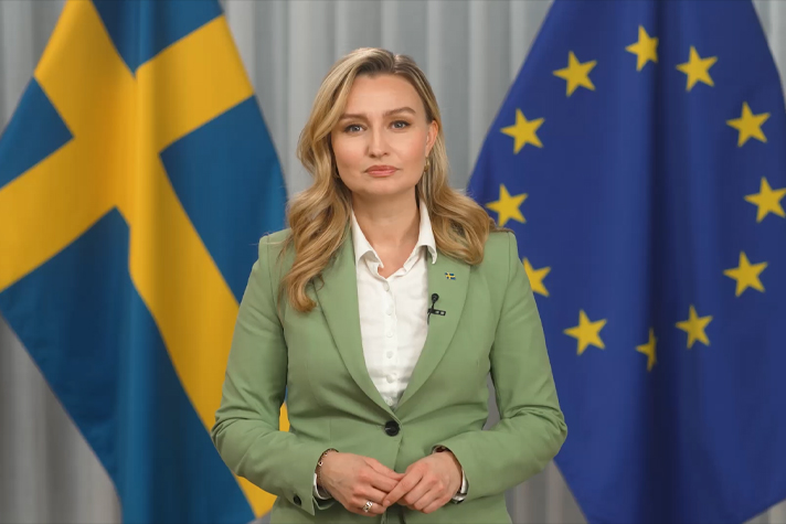 Bilden föreställer en kvinna framför en vit vägg. I bakgrunden synsflaggor.