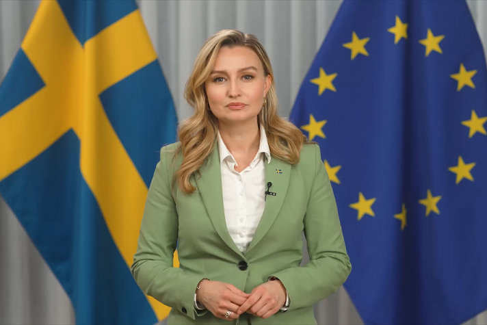 Bilden föreställer en kvinna framför en vit vägg. I bakgrunden synsflaggor.