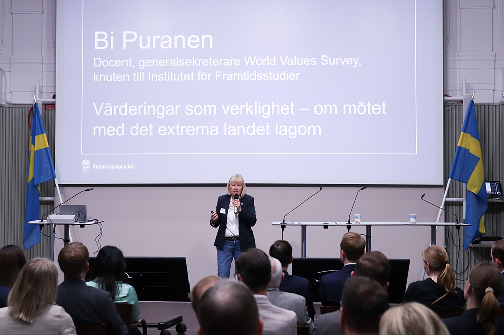 En kvinna talar i en mikrofon på en scen. I förgrunden syns huvuden på publiken. I bakgrunden syns en stor skärm med texten Bi Puranen, Värderingar som verklighet.