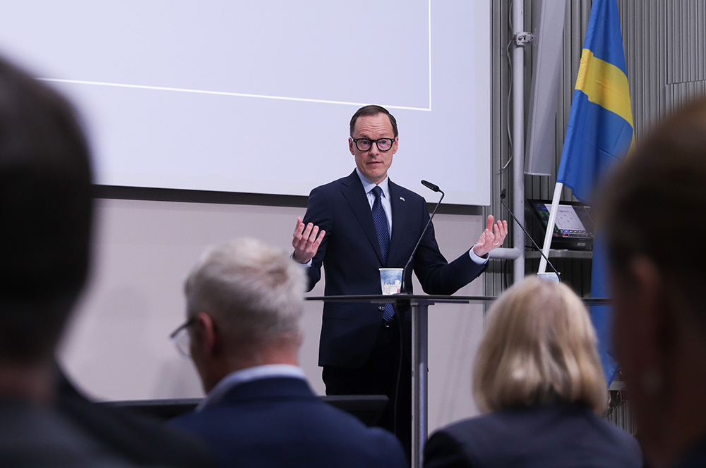 Mats Persson står på scenen och talar inför en publik. I förgrunden syns bakhuvuden på personer i publiken.