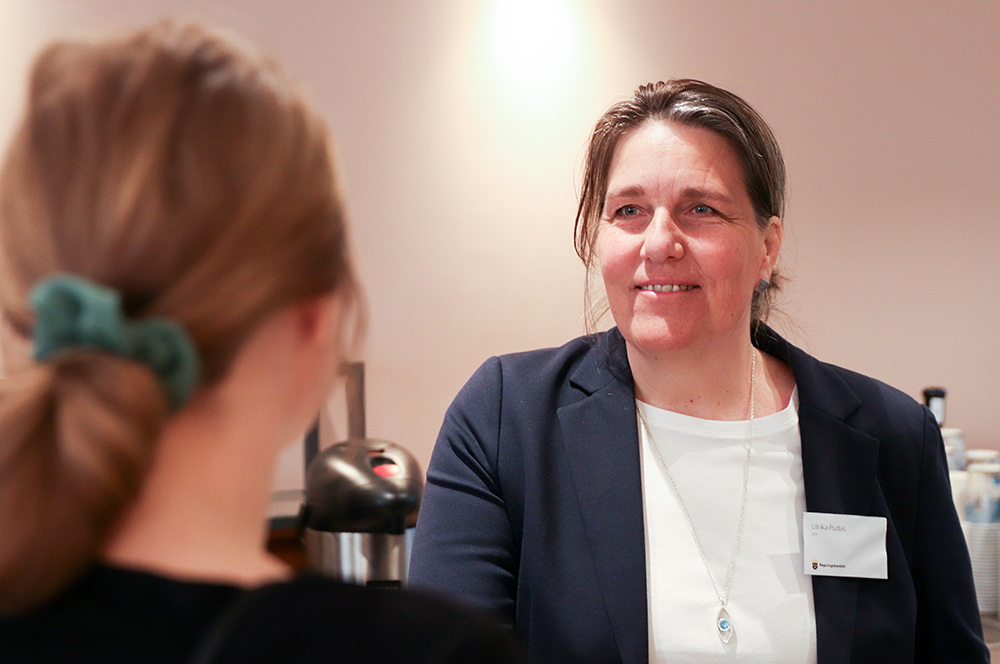 En kvinna ler och talar med en person som står med ryggen mot kameran.