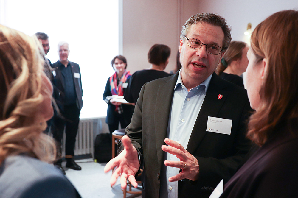 En man gestikulerar med händerna och talar med en kvinna. I bakgrunden syns några andra personer.