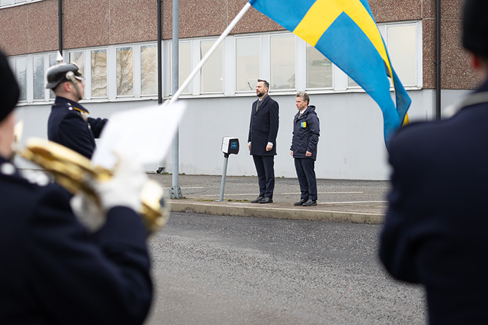 De polska och svenska försvarsministrarna står i givakt framför hedersvakten. De båda statsråden är i fokus under och bakom en svensk flagga som är ofokuserad. 