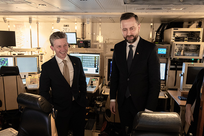 Pål Jonson och Władysław Kosiniak-Kamysz står i en mörk ledningscentral ombord en korvett. De båda ler. 