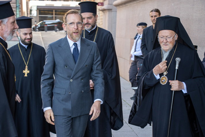 Socialministern möter Patriark Bartholomeus I utanför en stenbyggnad. Fotot är taget i stunden när de är på väg in i byggnaden.