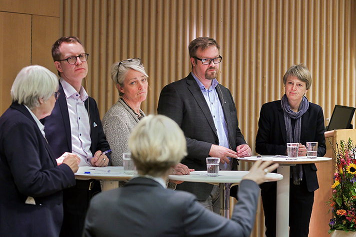 Fem personer som står på en scen bakom två bord och deltar i en paneldebatt. I förgrunden, en moderator.