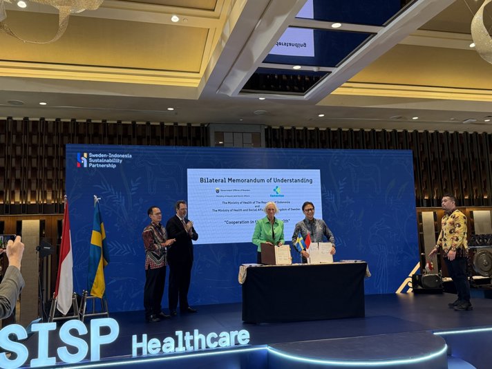 Två personer står på en scen och håller upp varsin pärm. På bordet framför dem står en svensk flagga och en indonesisk flagga. På skärmen bakom står det "Bilateral Memorandum of Understanding"