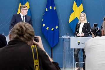 Gunnar Strömmer och Paulina Brandberg står vid mikrofoner framför fotografer i en presskonferenssal. Svenska flaggor och EU-flaggan i bakgrunden.