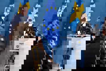 Gunnar Strömmer och Paulina Brandberg står vid mikrofoner framför fotografer i en presskonferenssal. Svenska flaggor och EU-flaggan i bakgrunden.