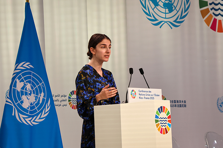 Ministern står i talarstolen intill FN-flaggan.