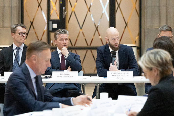 Fyra personer sitter vid ett bord, en talar i mikrofon. I förgrunden syns fler mötesdeltagare.