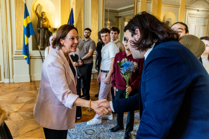 Två personer tar i hand. En grupp unga människor står bredvid. I bakgrunden skymtar två flaggor, en för Sverige och en för EU.