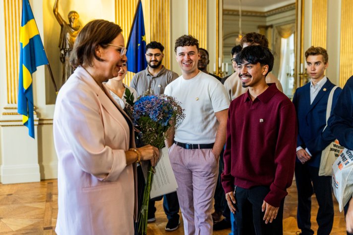 En ung man ger blommor till ministern. En grupp unga människor står bredvid. I bakgrunden skymtar två flaggor, en för Sverige och en för EU.
