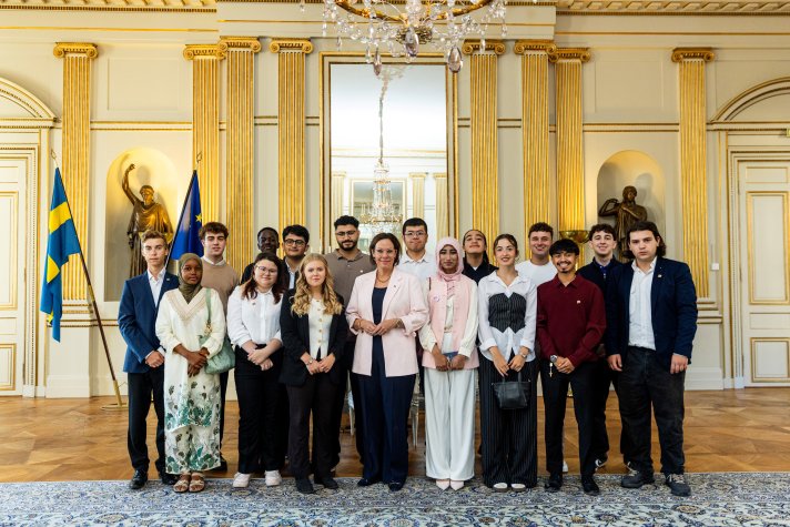 Utrikesministern och sexton unga pristagare står samlat för gruppfoto framför en vägg med gyllene dekorationer.
