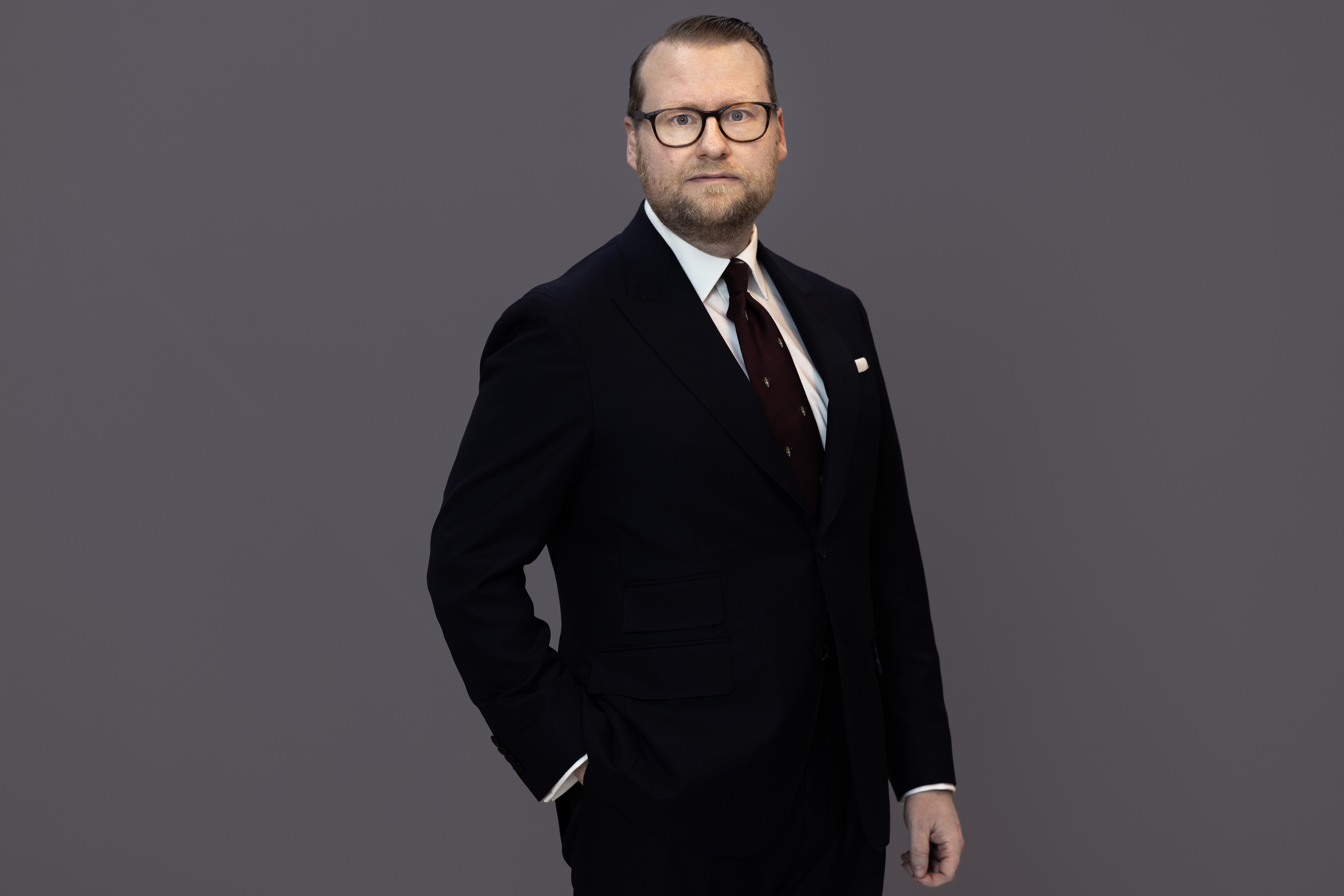 Statssekreterare Simon Westberg står framför en grå bakgrund, klädd i mörk kostym, vit skjorta och mörk slips. Han bär glasögon och ser rakt mot kameran med ena handen i fickan.