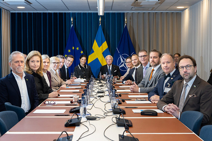 14 personer sitter runt ett stor mötesbord, formell miljö. I bakgrunden syns EU-flaggan, Svenska flaggan och Nato-flaggan.