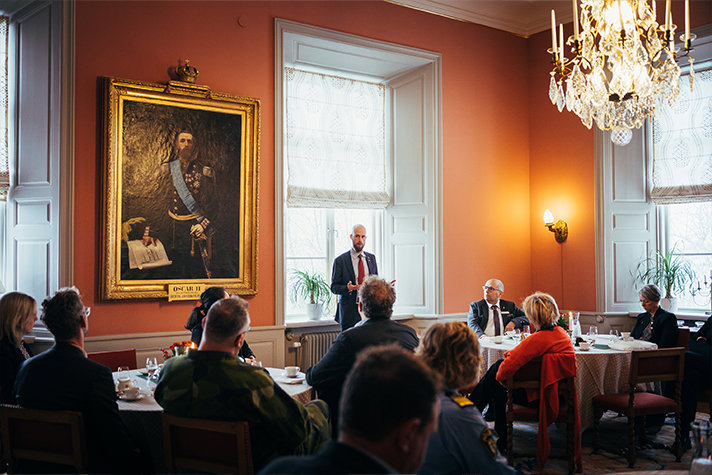 Minister för civilt försvar Carl-Oskar Bohlin håller ett anförande inne i ett rum på landshövdingens residens i Linköping. På bilden syns personer som sitter runt bord och lyssnar.