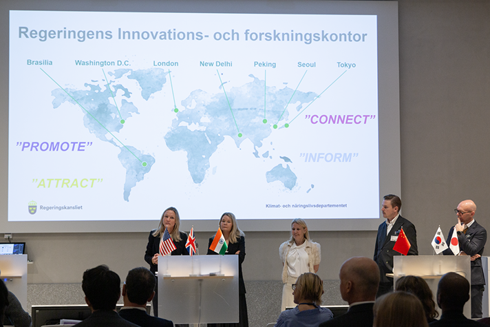 Fem personer som presenterar vad de som regeringens innovations- och forskningsråd arbetar med. De står i en stor aula framför en en världskarta där de olika kontoren är utmärkta.