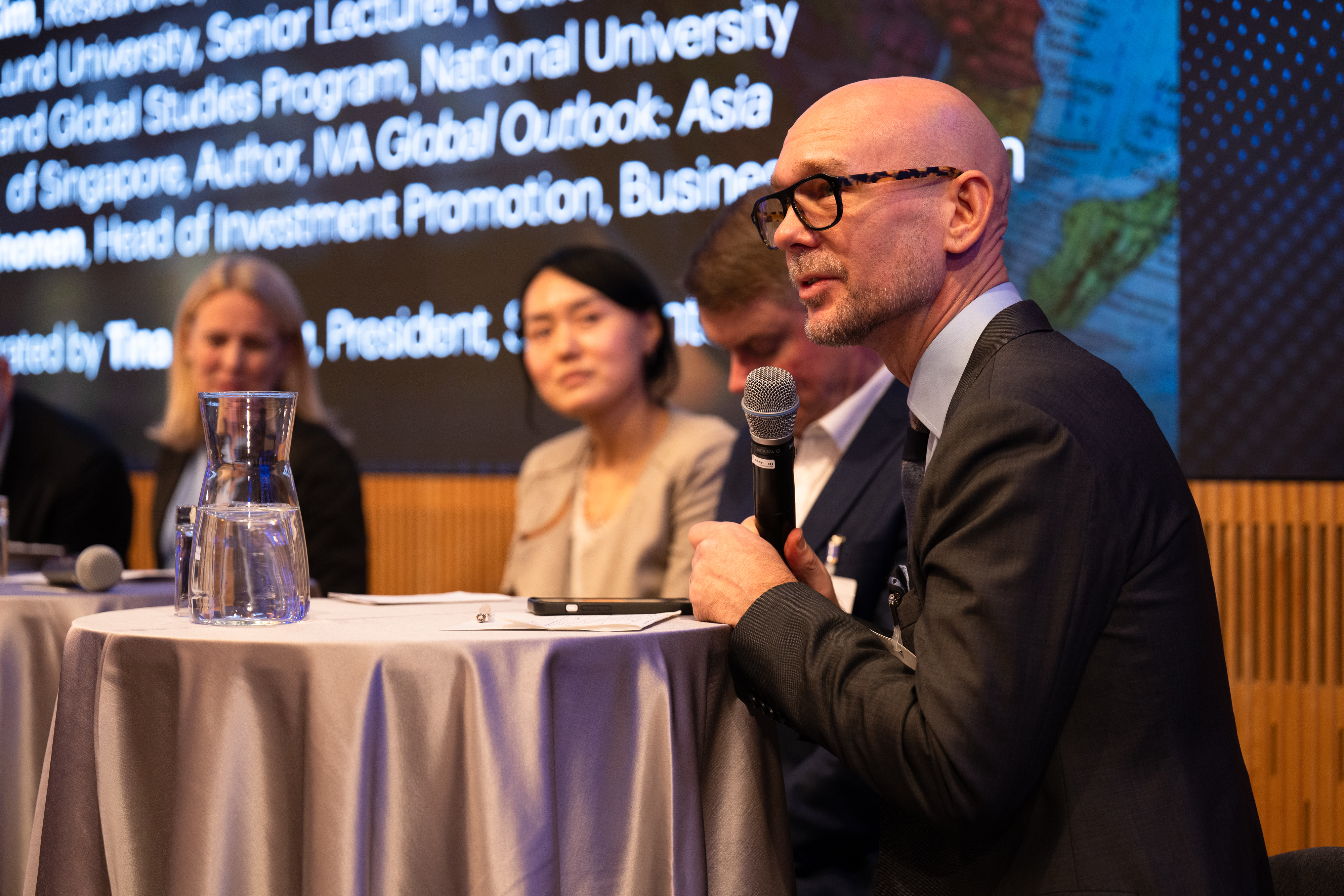 En man med glasögon talar i en mikrofon, och i bakgrunden syns flera andra personer som deltar i ett panelsamtal.