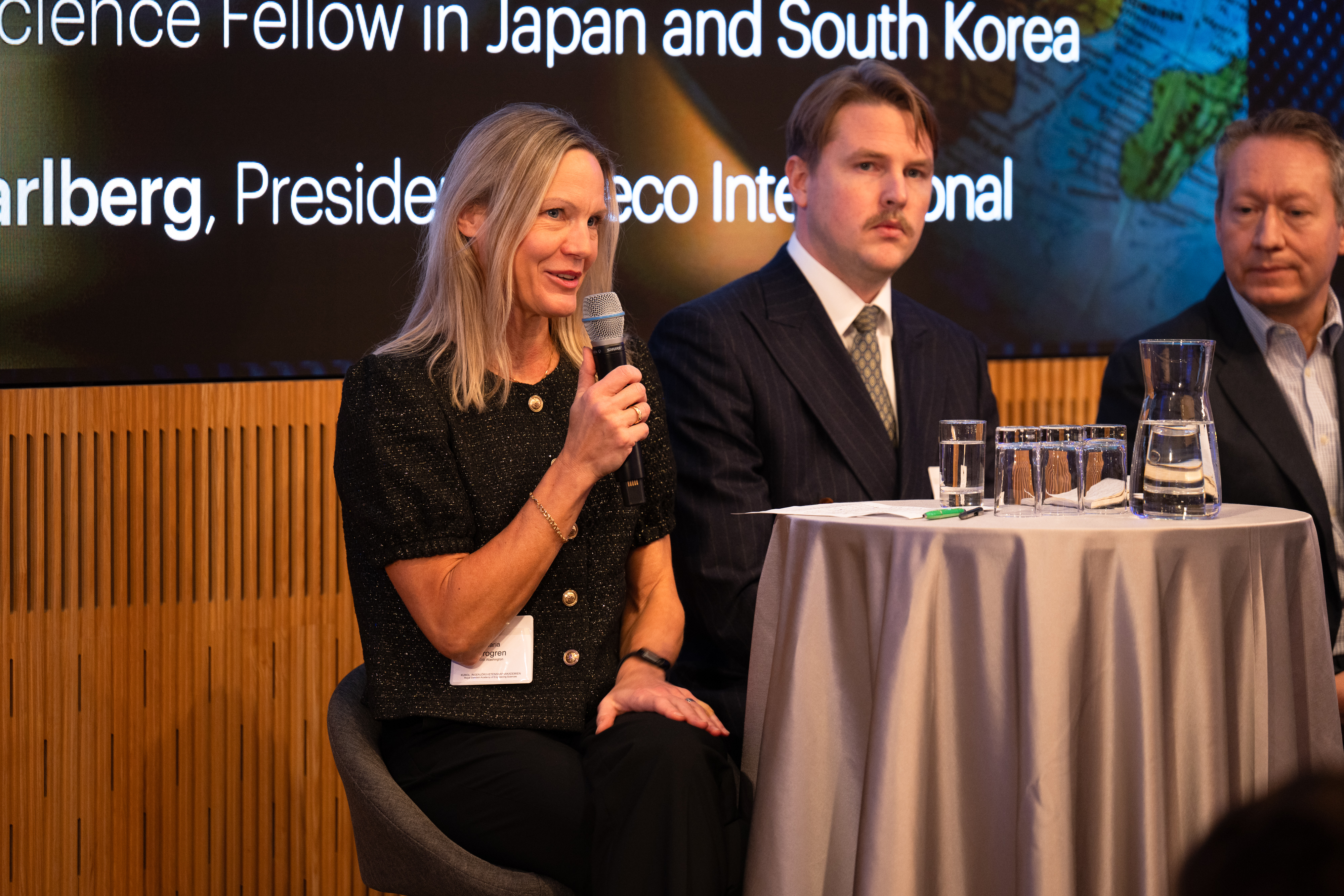 En kvinna talar i en mikrofon, och vid hennes sida sitter två män i en soffa.