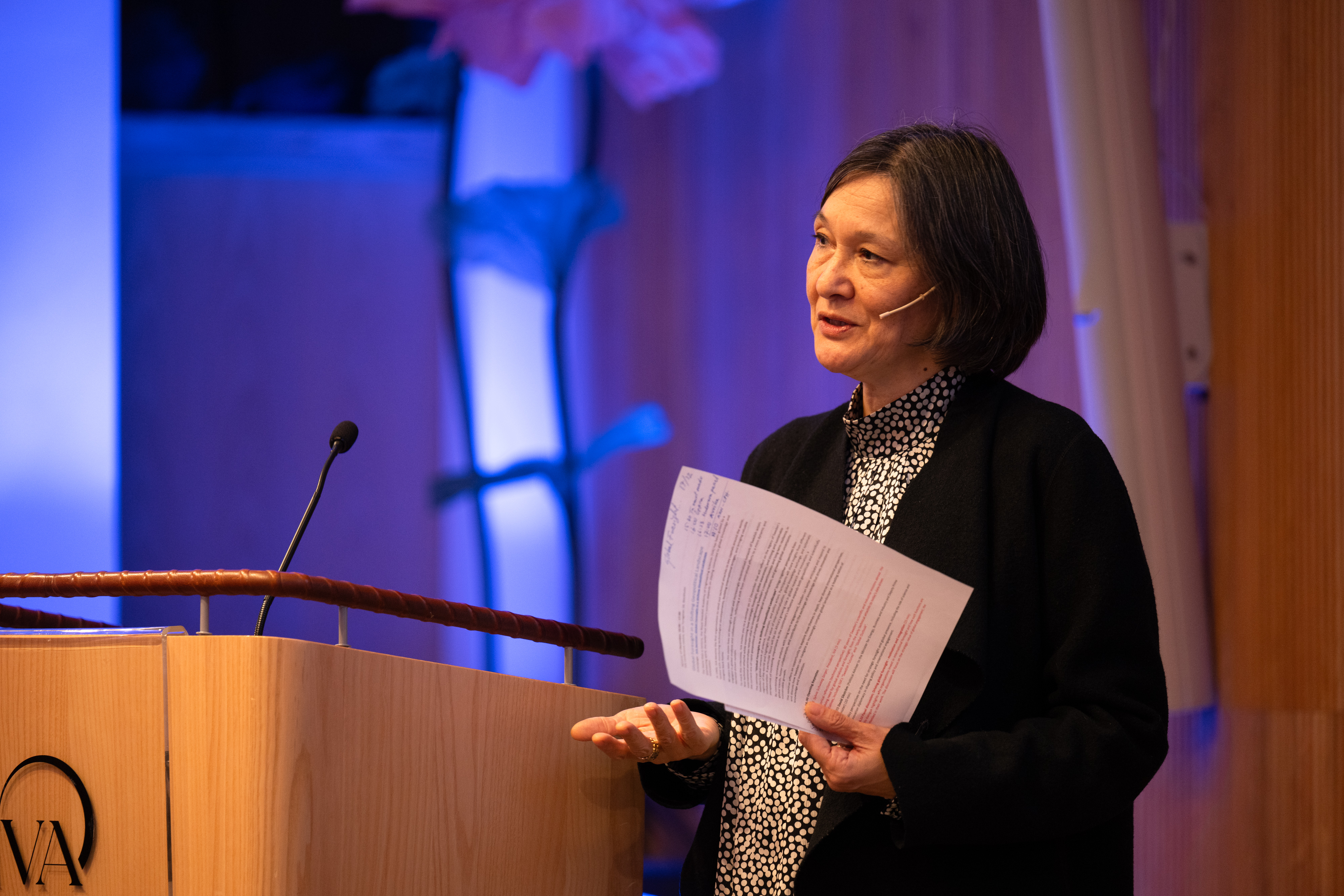 En kvinna med mörkt hår håller ett papper framför sig, med blå bakgrund bakom henne.