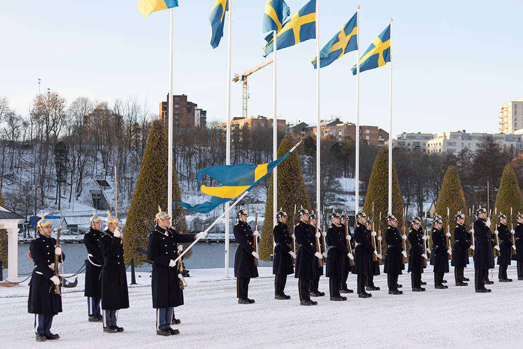 Hedersvakt står i givakt i två rader. En svensk flagg hålls fram av en soldat medans andra flaggor vajar i vinden bakom hedersvakten