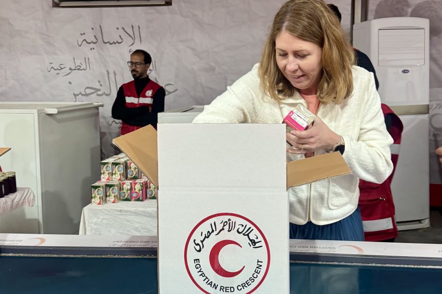 Sjukvårdsminister Elisabet Lann packar humanitära förnödenheter i en kartong märkt med Egyptian Red Crescent. I bakgrunden syns personal i röda västar och staplar av hjälpprodukter.