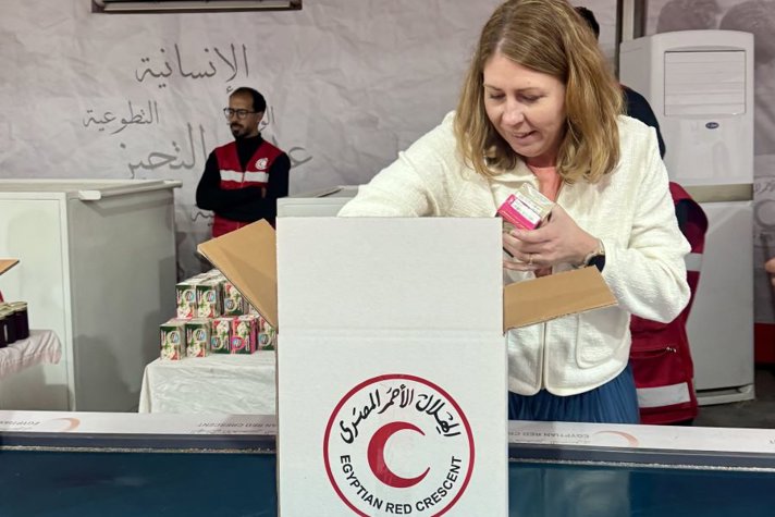 Sjukvårdsminister Elisabet Lann packar humanitära förnödenheter i en kartong märkt med Egyptian Red Crescent. I bakgrunden syns personal i röda västar och staplar av hjälpprodukter.