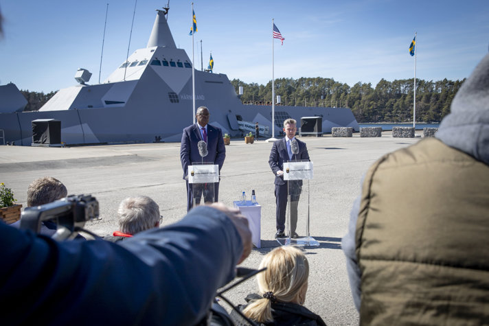 Försvarsminister Lloyd Austin och försvarsminister Pål Jonson under en pressträff. I bakgrunden syns Visby-korvetten Härnösand bakom två flaggor. En svensk och en amerikansk. 