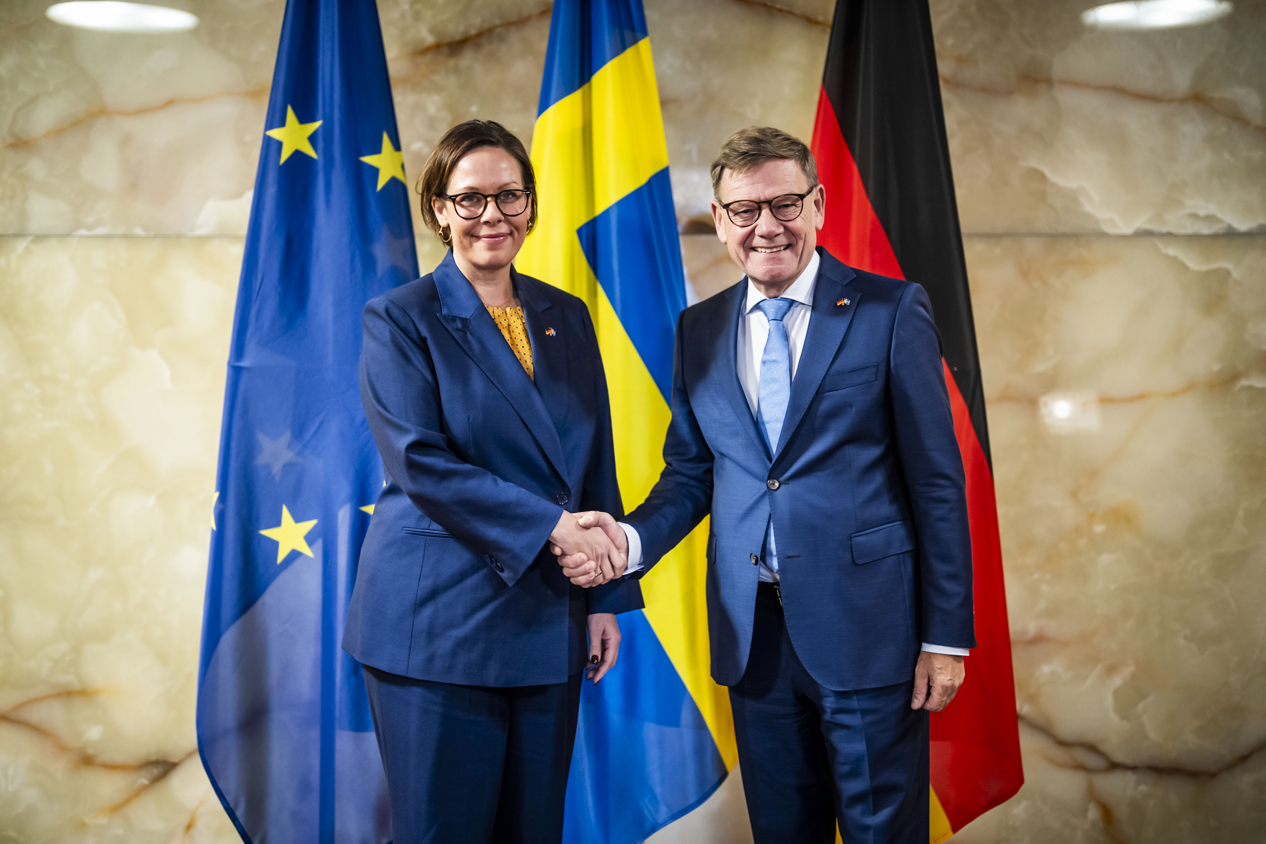 Maria Malmer Stenergard och Johann Wadepuhl skakar hand framför tysk flagga, svensk flagga och EU-flagga. 