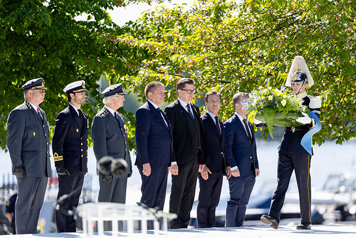 Statsråd, H.M. konungen och överbefälhavaren m.fl. står på rad på ett podium bredvid soldater som håller kransar.