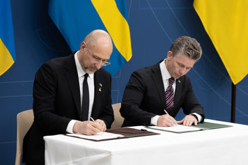 Två män i kostym sitter sida vid sida vid ett bord och signerar dokument. I bakgrunder skymtar svenska och ukrainska flaggor.