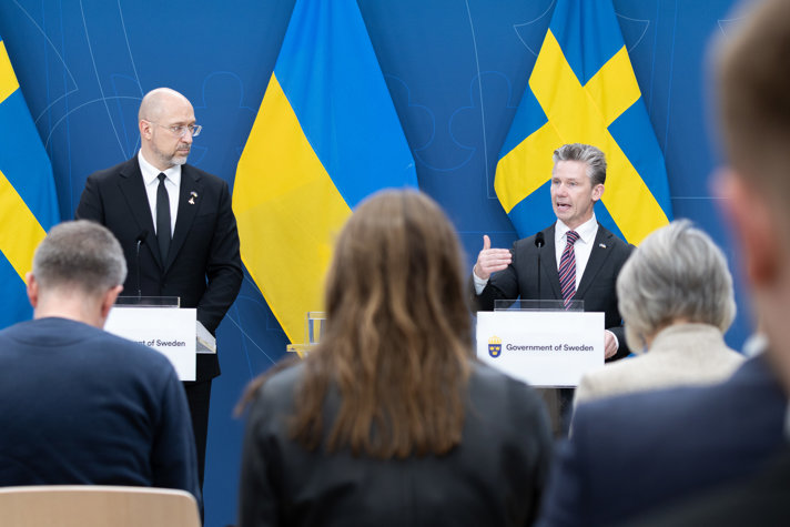 Två män i kostym står vid varsin talarstol och håller pressträff. I bakgrunden skymtar svenska och ukrainska flaggor. I förgrunden syns publik bakifrån som lyssnar på talarna.