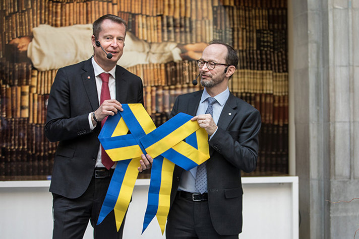 De två statsråden håller upp en rosett i gult och blått som ska symbolisera ihopknytning av frågorna i det nya departementet.