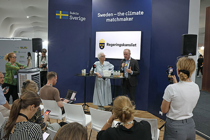 De två personerna står vid ett ståbord. Bakom dem en skärmvägg med texten Sweden the Cliamte Matchmaker. Reportrar syns i förgrunden.